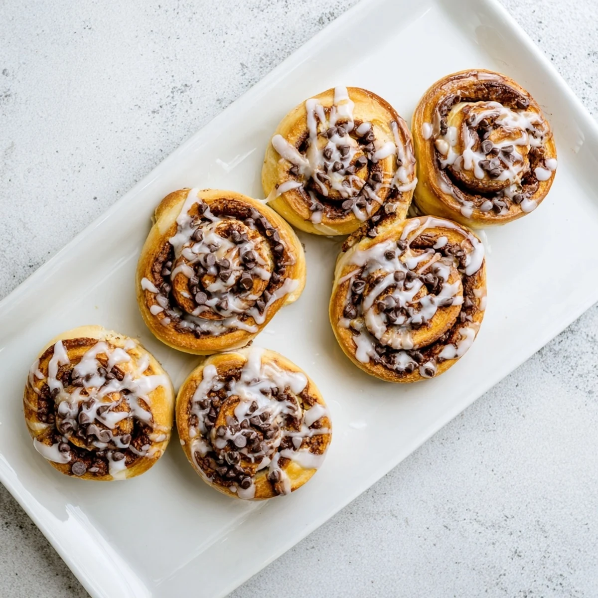 Warm, freshly baked Nutella Chocolate Chip Rolls sit in a pan, their golden-brown spirals glistening with a sweet glaze and dotted with melty chocolate chips.