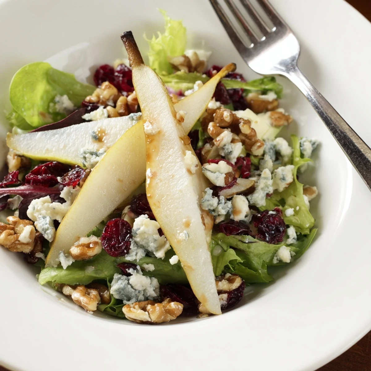 Fresh sliced pears and crumbled Gorgonzola on crisp mixed greens with toasted walnuts, drizzled with honey-balsamic vinaigrette in a white serving bowl.