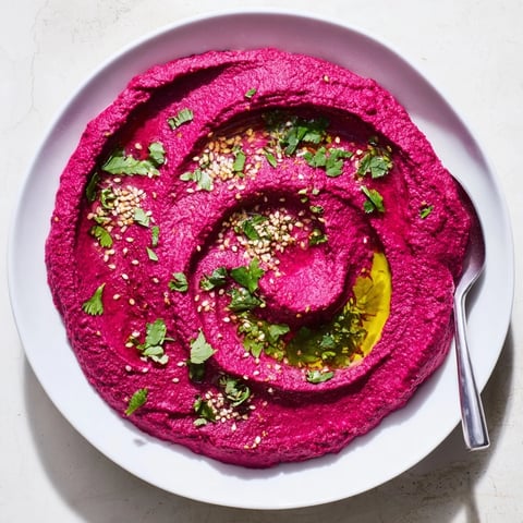 Creamy roasted beet hummus topped with olive oil in a rustic serving bowl, surrounded by fresh pita chips and vegetable sticks for dipping.