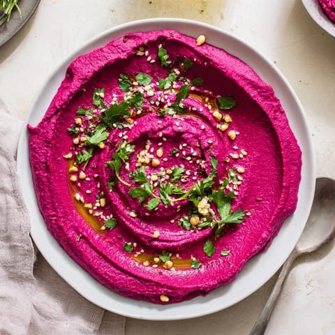 A vibrant bowl of roasted beet hummus garnished with parsley and sesame seeds, next to colorful sliced veggies for a healthy snack platter.